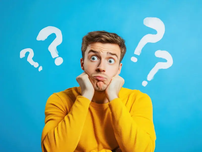 Why Every Website Needs an FAQ Page - this image has abright blue background with a man in a bright yellow jumper sat with elbows on the desk and his chin resting in his hands, he looks confused and there are white question marks either side of him